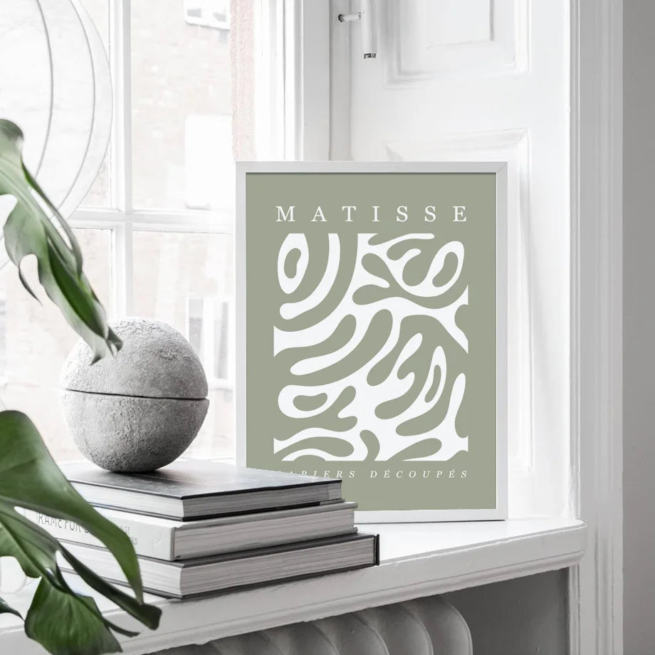 Framed artwork with 'Matisse' text and abstract design on a shelf with books and a plant.