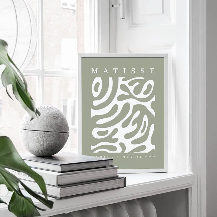 Framed artwork with 'Matisse' text and abstract design on a shelf with books and a plant.